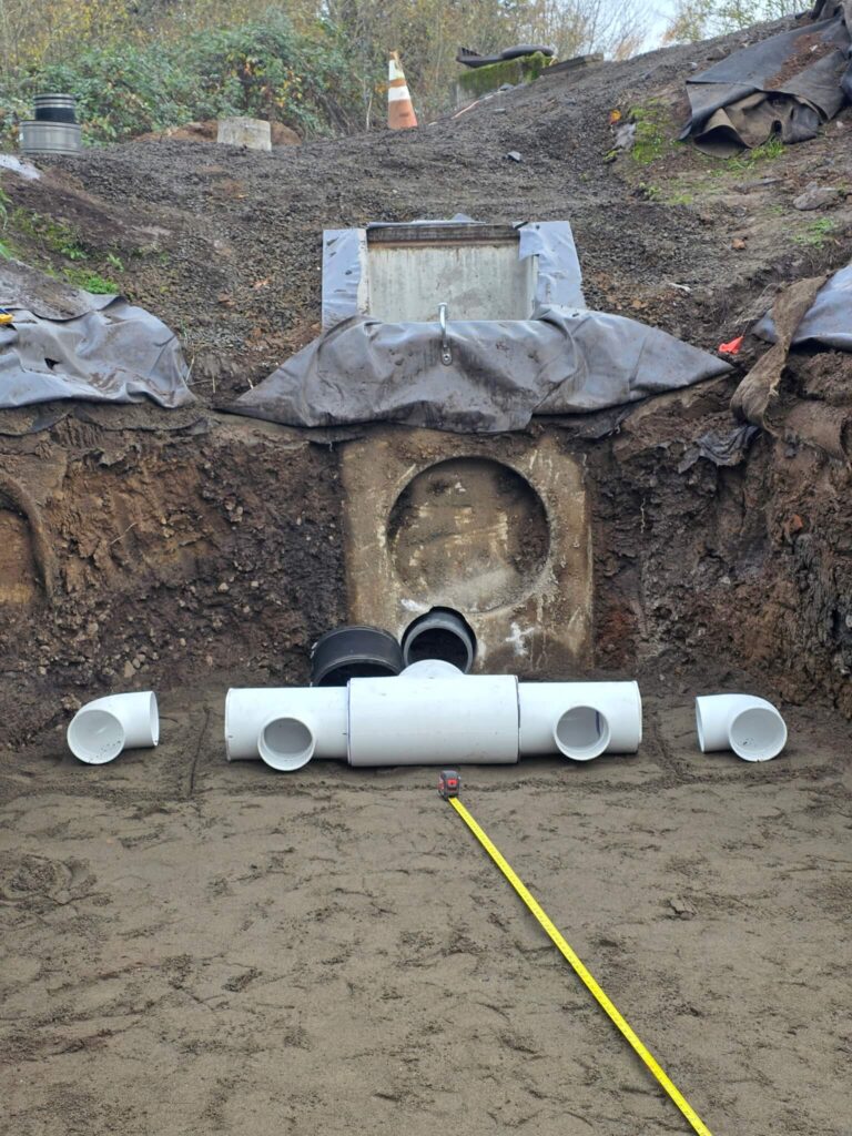 Excavated detention pond showing core-drilled concrete outlet structure and PVC collection pipe header staged for connection, during biochar biofilter installation at Oregon waste transfer facility